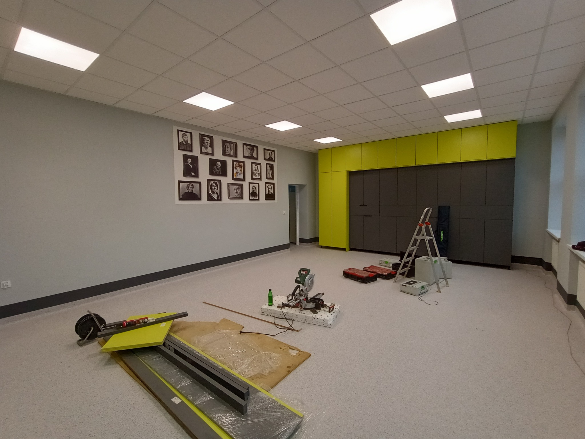 Wnętrze biura w trakcie remontu: widoczne narzędzia, drabina, panele podłogowe, ściany pomalowane na szaro, szafki w kolorze limonkowym i ciemnoszarym oraz portrety na ścianie