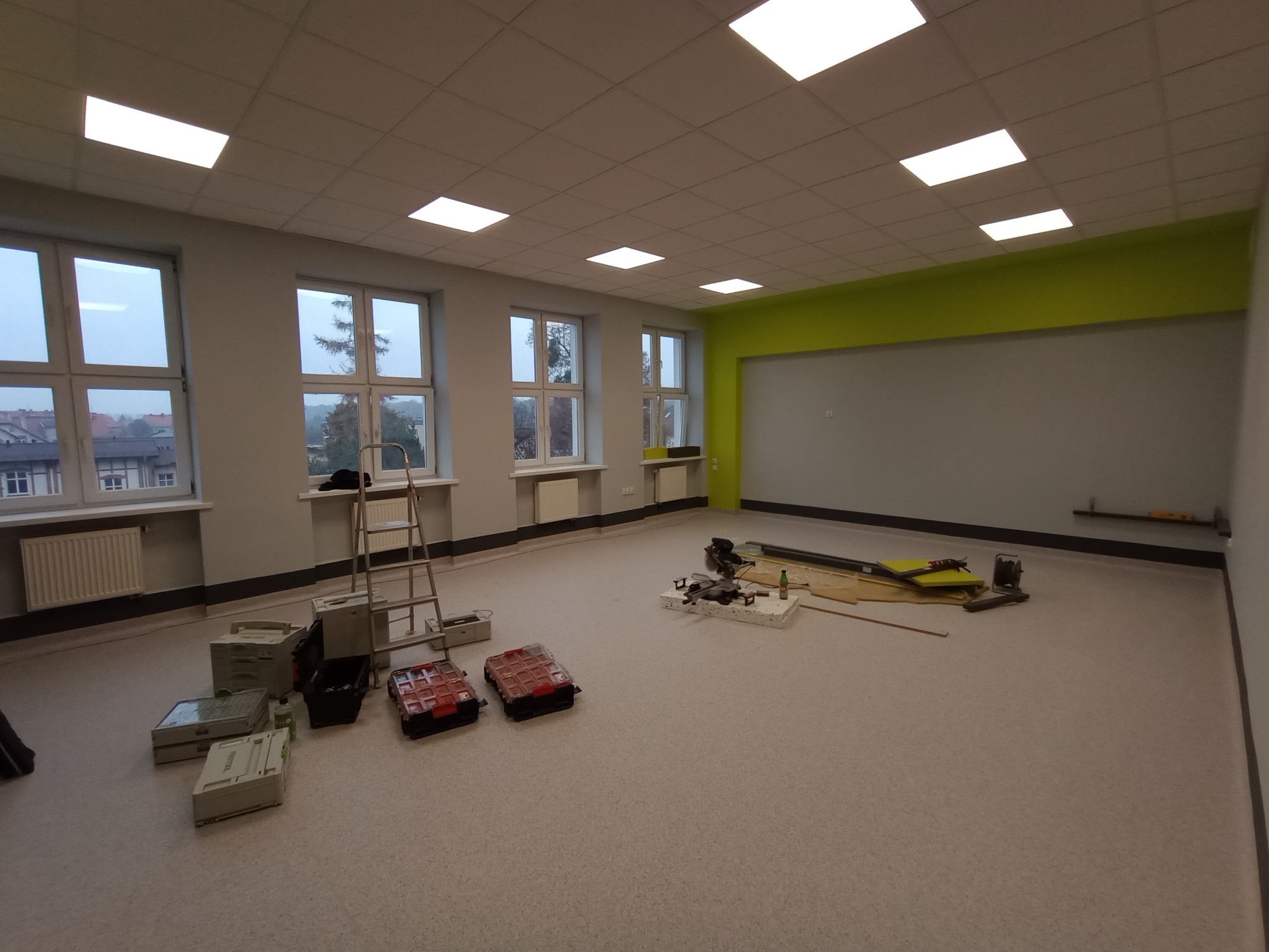 Przestronne pomieszczenie w trakcie prac wykończeniowych: ściany pomalowane na szaro z zielonym akcentem, narzędzia i skrzynki narzędziowe na podłodze, drabina oparta o ścianę.