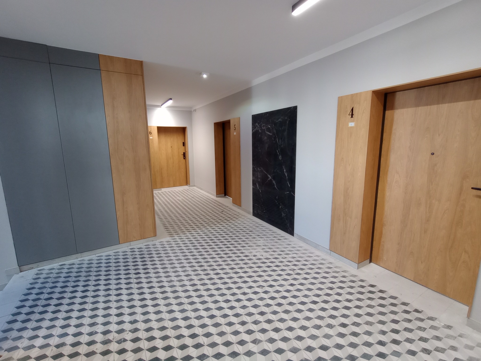 Korytarz w nowym budownictwie z wzorzystą podłogą, drewnianymi drzwiami z numerami mieszkań i panelami ściennymi w odcieniach szarości i drewna.