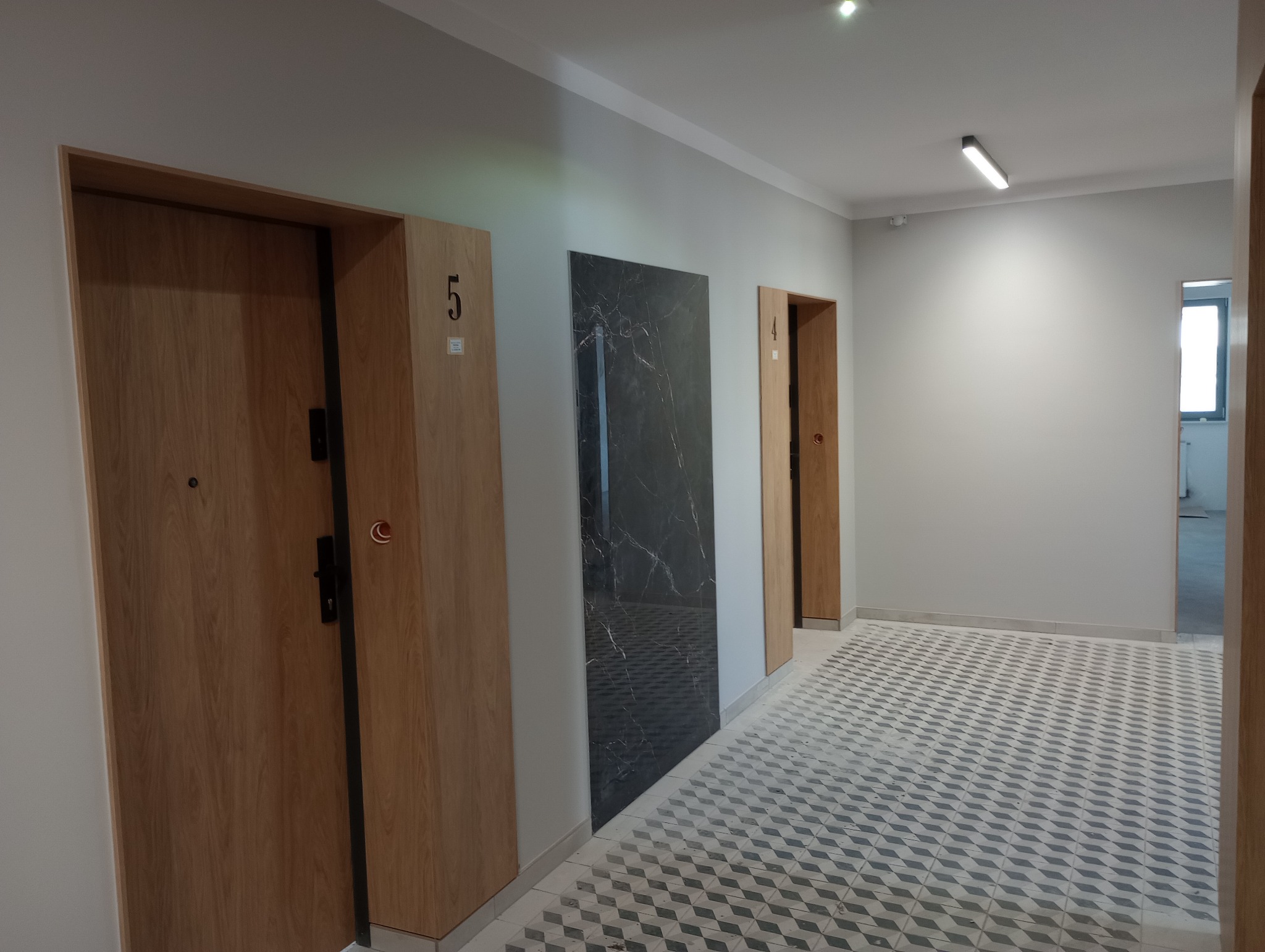 Korytarz w nowym budownictwie z trzema drzwiami o numerach 4 i 5, wykończony płytkami podłogowymi w geometryczny wzór oraz z czarną płytą dekoracyjną na ścianie.