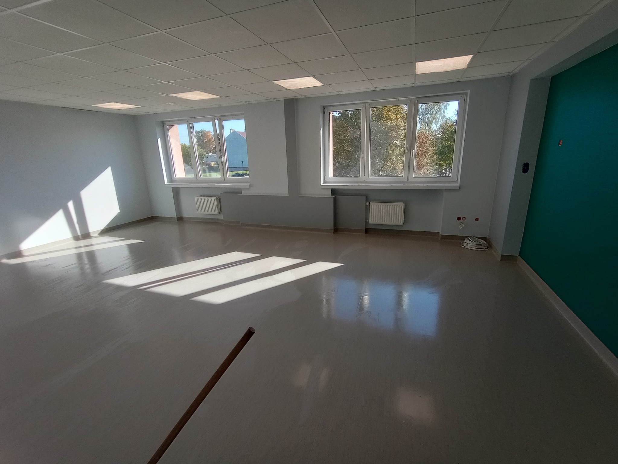 Pomalowane, jasne pomieszczenie biurowe z dwoma oknami, grzejnikami i częściowo pomalowaną na zielono ścianą, widoczne kable na podłodze i cień mopa.