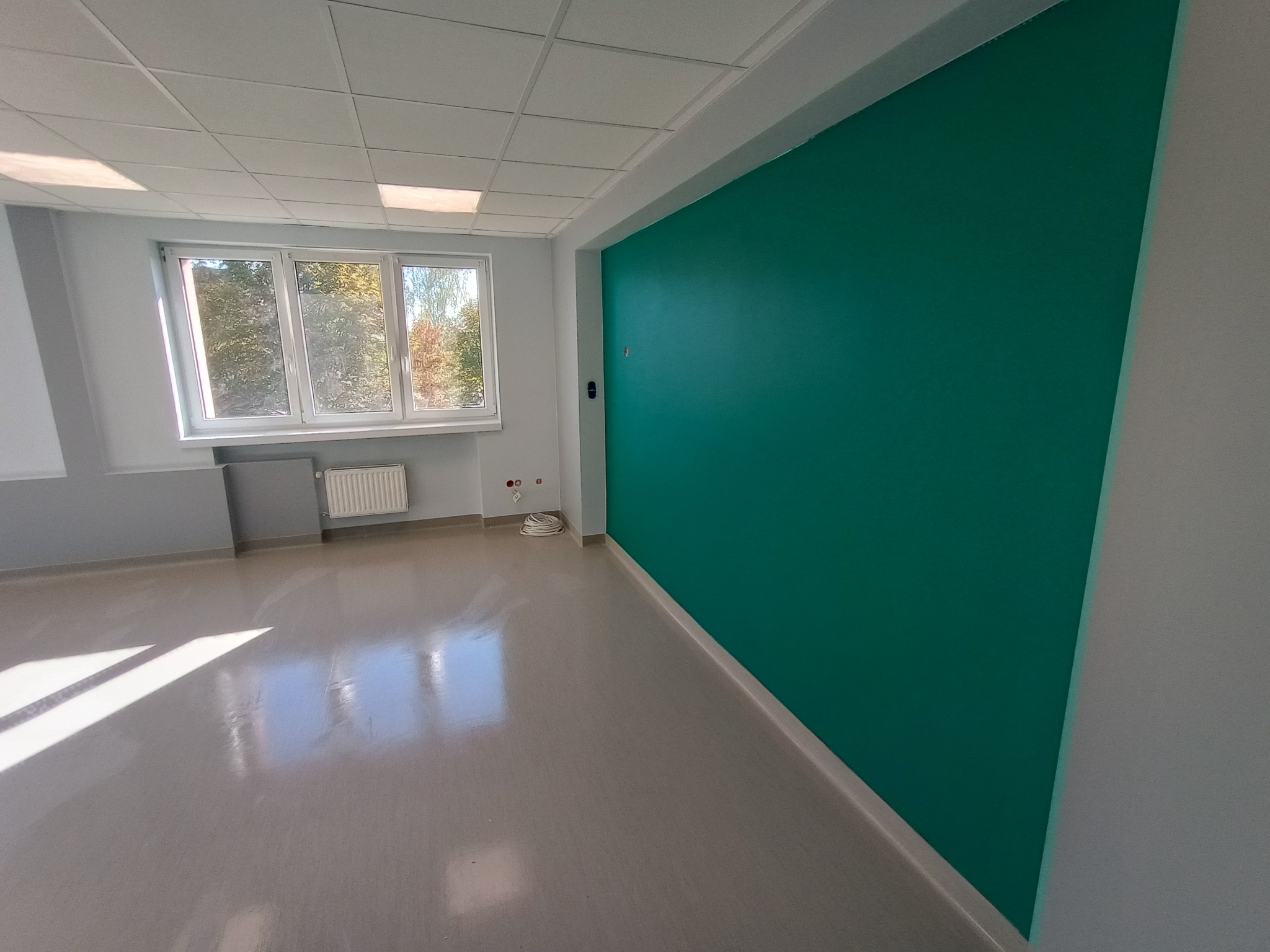 Pomalowane na turkusowo ściany w pustym, jasnym pomieszczeniu biurowym z oknem i grzejnikiem, widoczne gniazdka elektryczne.