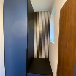 Niebiesko-czarna szafa z siedziskiem w przedpokoju, z czarno-białą ścianą w tle i drewnianymi drzwiami po prawej stronie.