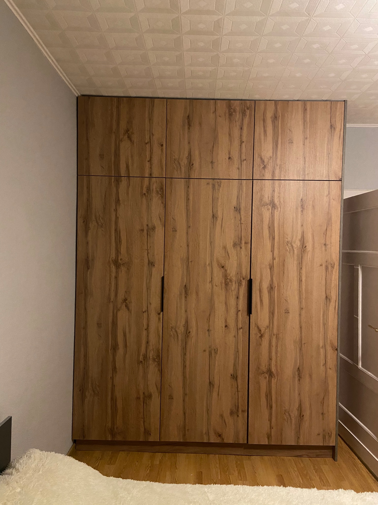 Wysoka, zabudowana szafa z drewnopodobnymi frontami i czarnymi, pionowymi uchwytami, sięgająca sufitu z kasetonami, widziana w rogu pokoju.