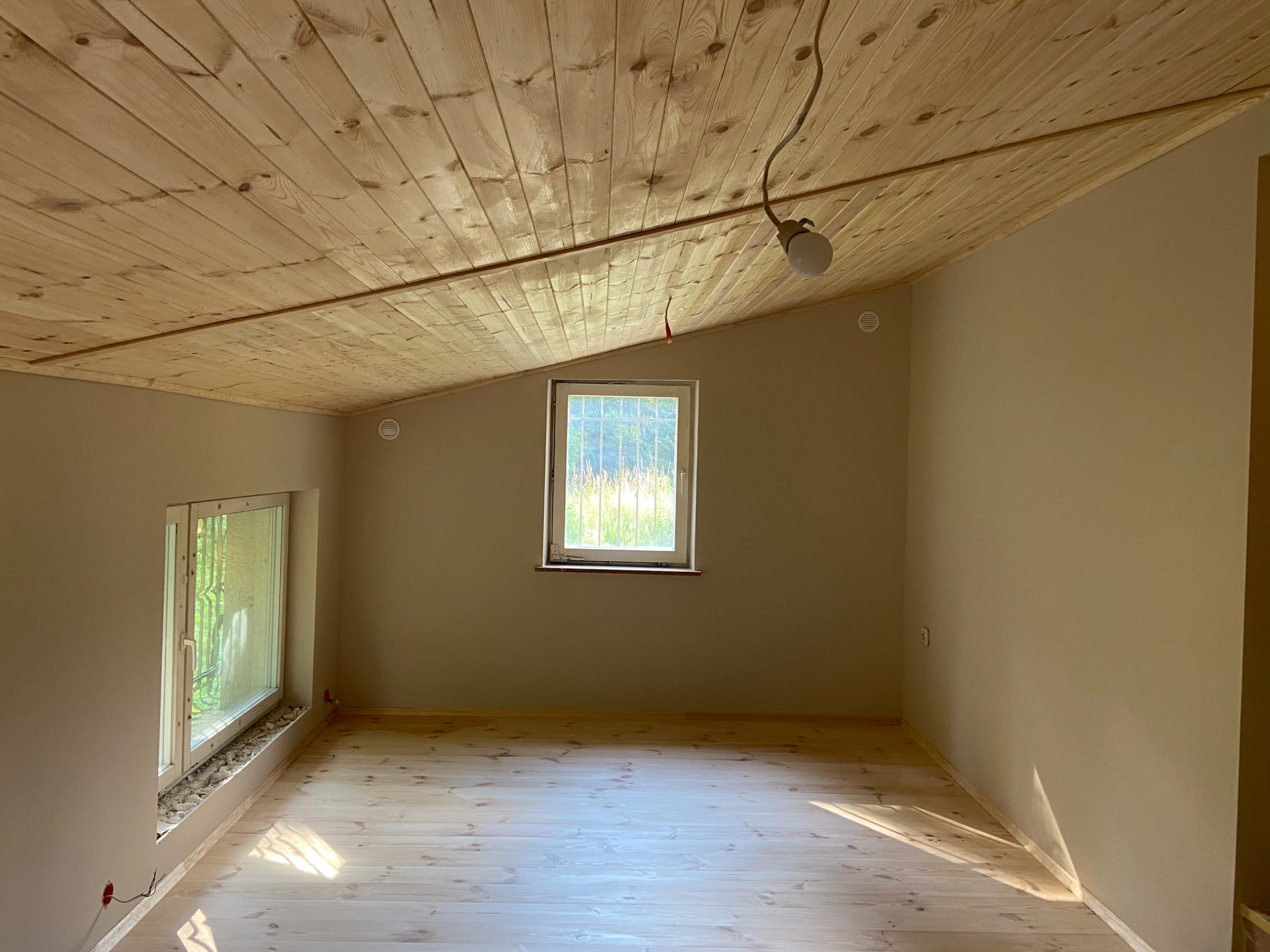 Poddasze z drewnianą boazerią na suficie i jasnymi ścianami, widoczne okna i niedokończone instalacje elektryczne.