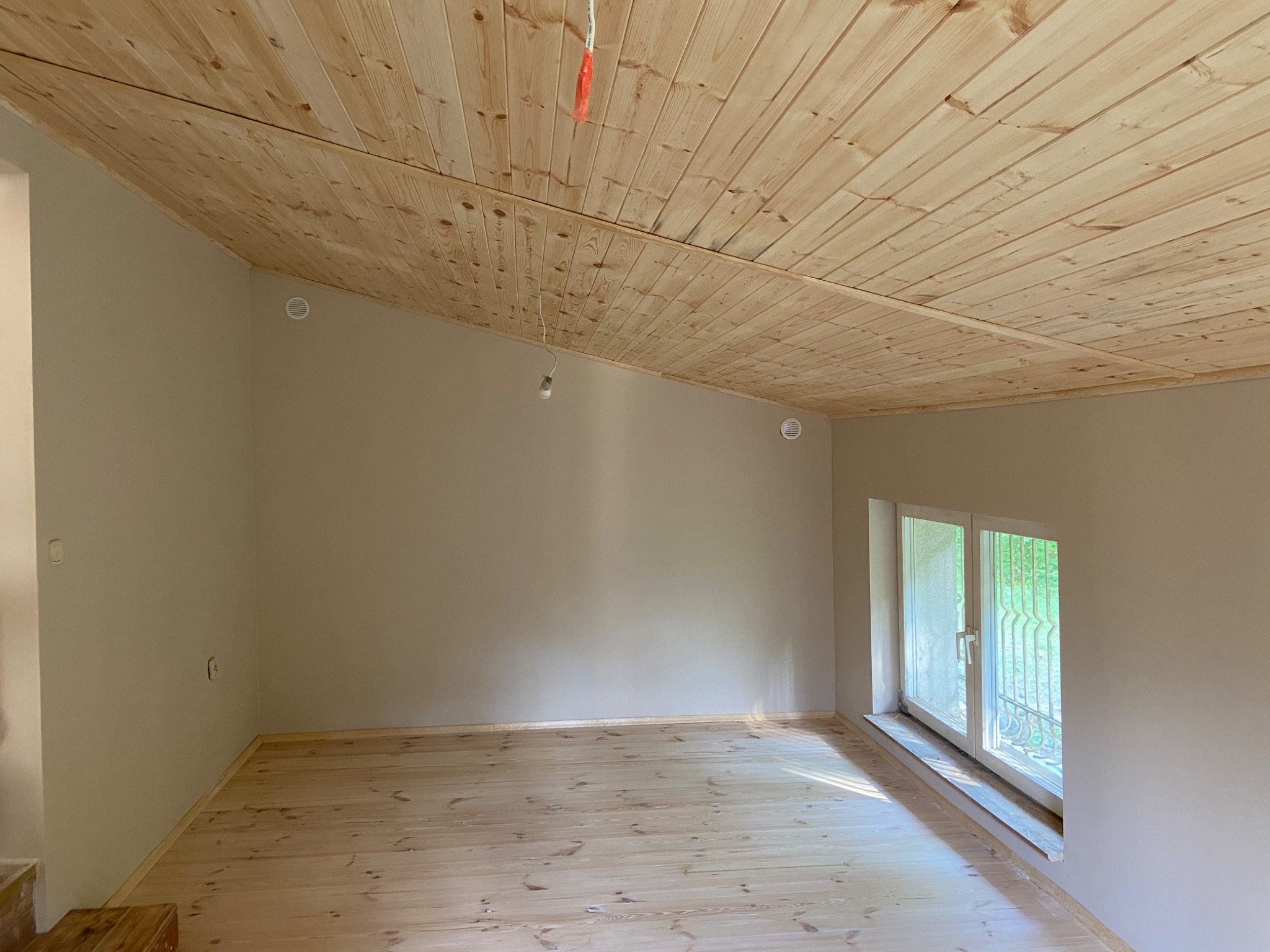 Pokój po remoncie: drewniana podłoga i sufit z desek, ściany pomalowane na beżowo, widoczne okno i niedokończone oświetlenie sufitowe.