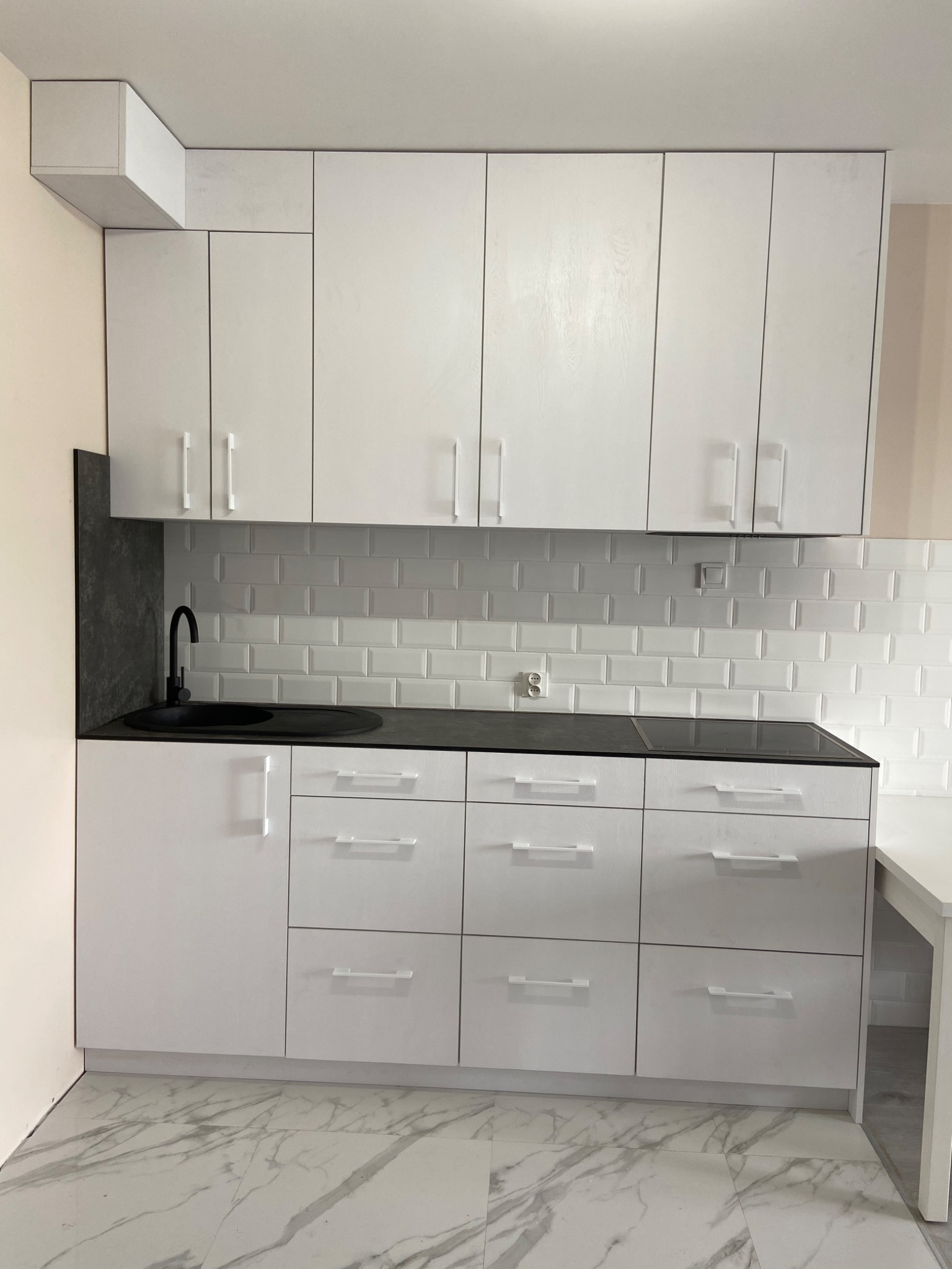 Biała zabudowa kuchenna z czarnym blatem i okrągłym zlewem, uzupełniona białymi płytkami w stylu metro na ścianie i marmurową podłogą.
