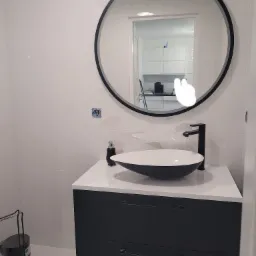 Nowoczesna łazienka z okrągłym lustrem w czarnej ramie, białą umywalką nablatową w kształcie misy i czarną armaturą na szafce z ciemnymi szufladami, białe płytki z delikatnym wzorem na ścianie.