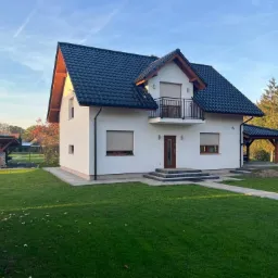 Dwukondygnacyjny dom z białymi ścianami, ciemnym dachem i drewnianymi elementami wykończeniowymi, otoczony zielonym trawnikiem i drzewami w jesiennej scenerii.