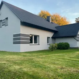 Szary dom jednorodzinny z czarnym dachem i dekoracyjnymi pasami na elewacji, otoczony zielonym trawnikiem w słoneczny dzień.