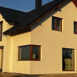 Jasna elewacja domu jednorodzinnego z ciemną dach&oacute;wką i brązowymi ramami okiennymi, widoczne rolki materiału izolacyjnego za oknem, ciepłe światło popołudniowe.