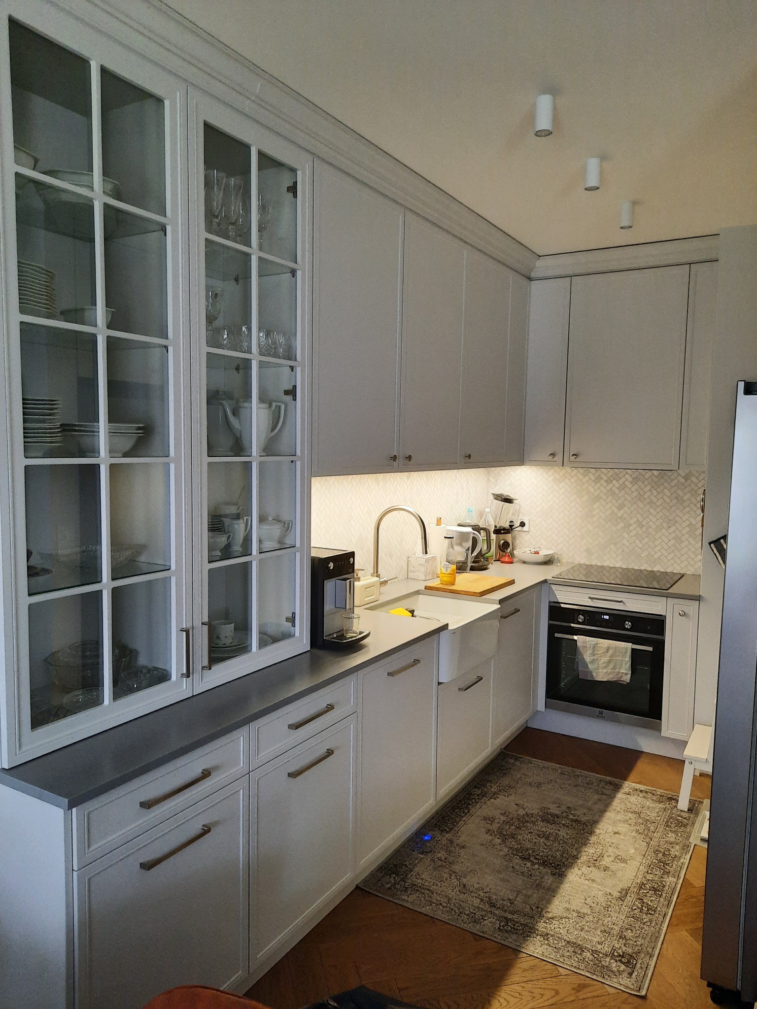 Jasna kuchnia z szarymi blatami, przeszklona szafka z zastawą, biały zlew farmerski i mozaika rybia łuska na ścianie.