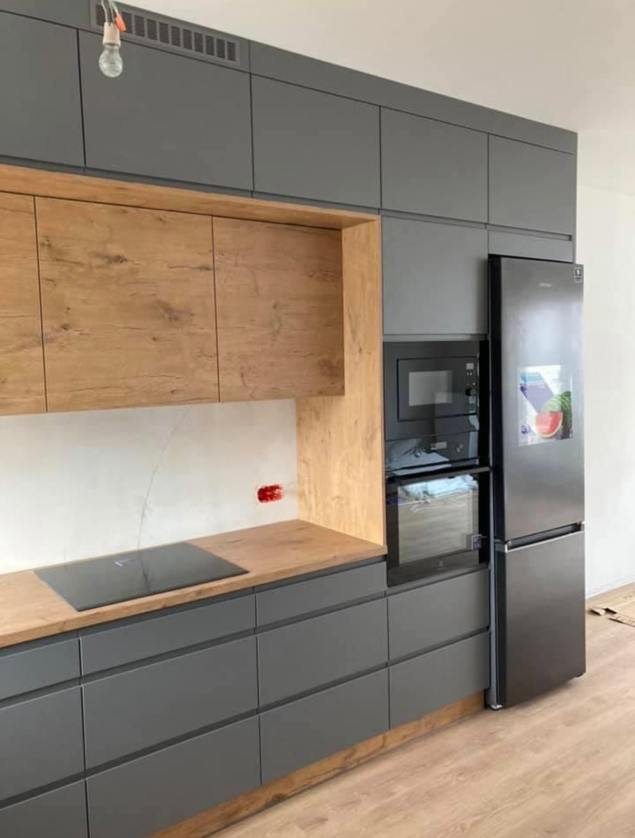 Nowoczesna kuchnia z szarymi szafkami, drewnianymi blatami i okładziną, czarnym sprzętem AGD (piekarnik i mikrofalówka w zabudowie, lodówka side-by-side) oraz płytą indukcyjną.