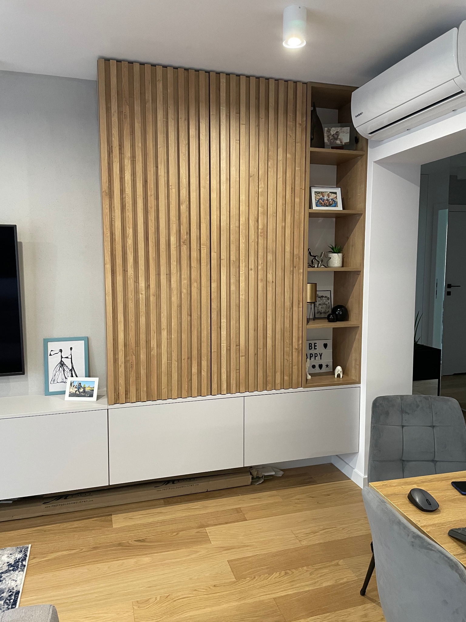 Nowoczesna szafa w salonie z drewnianymi lamelami na frontach i otwartymi półkami z dekoracjami, białe szafki dolne, widoczny klimatyzator i fragment stołu z krzesłami.