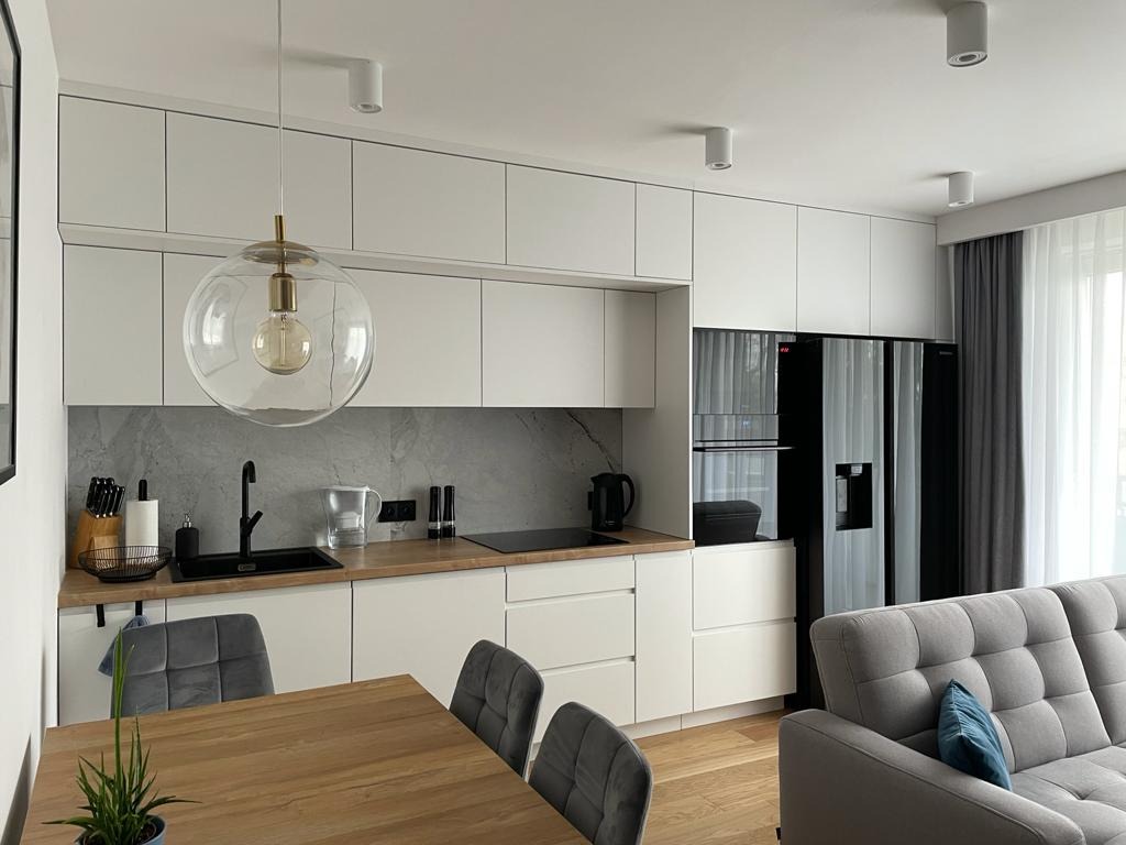 Nowoczesna kuchnia z białymi szafkami, drewnianym blatem i szarą marmurową ścianą, oświetlona kulistą lampą wiszącą nad drewnianym stołem z szarymi krzesłami, widok na czarną lodówkę i szarą kanapę.