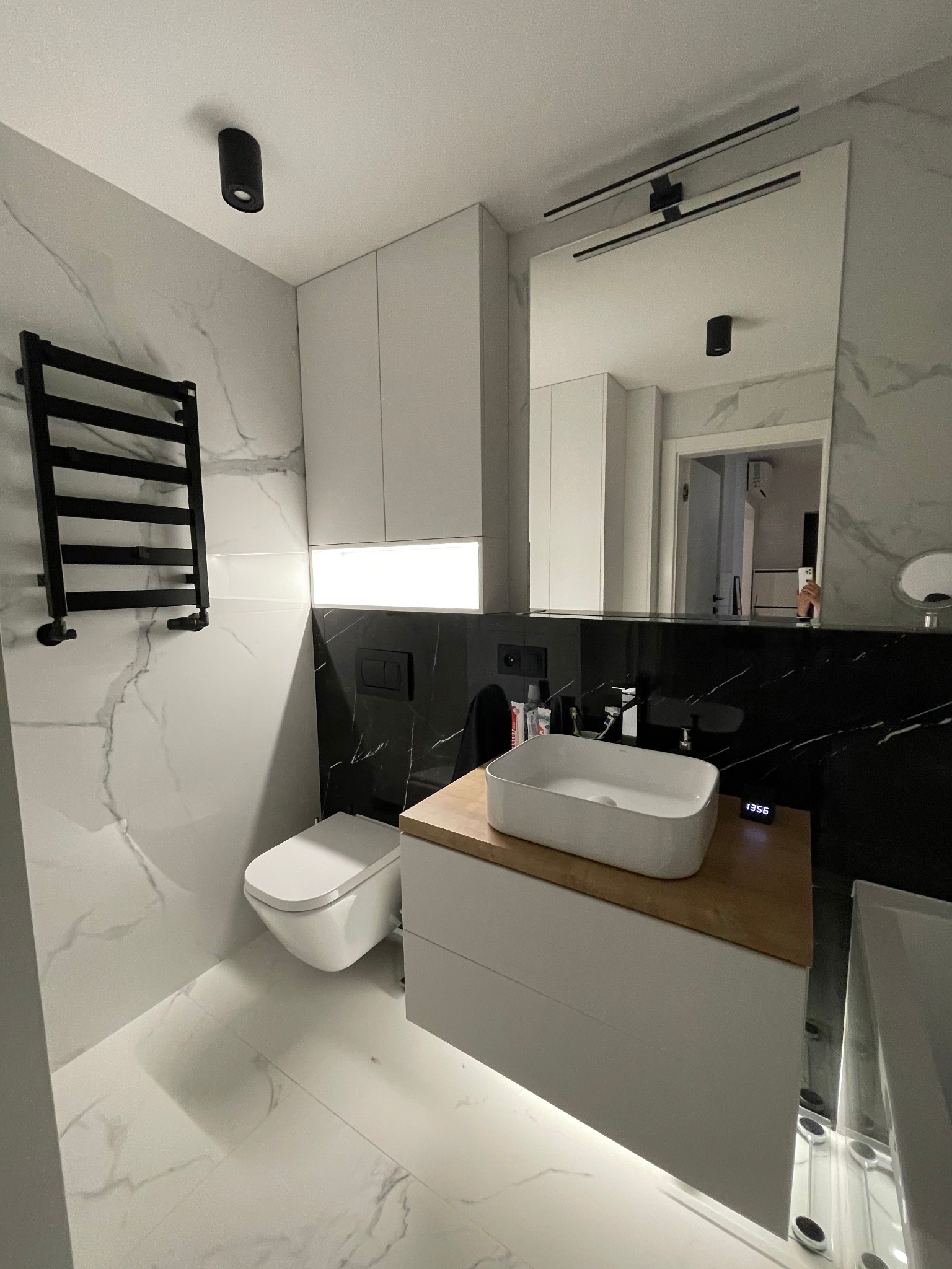 Nowoczesna łazienka z marmurowymi płytkami, wiszącą toaletą, grzejnikiem drabinkowym i umywalką nablatową na drewnianym blacie, oświetlona lampami sufitowymi i listwą LED nad szafką.