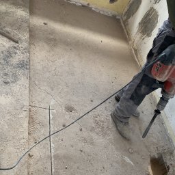 Tomasz Gałązka - Pracownik w Świdwinie używa młota pneumatycznego do skuwania posadzki w pomieszczeniu przed remontem, widoczne ślady po instalacjach na ścianach i resztki starej farby.