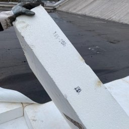 Tomasz Gałązka - Ocieplenie dachu: pracownik w rękawicy trzyma białą płytę styropianową z nadrukowanym numerem na tle płaskiego, bitumicznego dachu z widocznymi fragmentami ocieplenia i elementami konstrukcyjnymi.