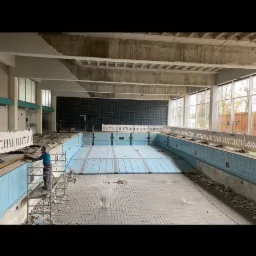 Pracownik w niebieskiej koszuli na rusztowaniu, usuwa płytki ze ściany nieczynnego basenu, widoczne gruz i resztki płytek na podłodze.