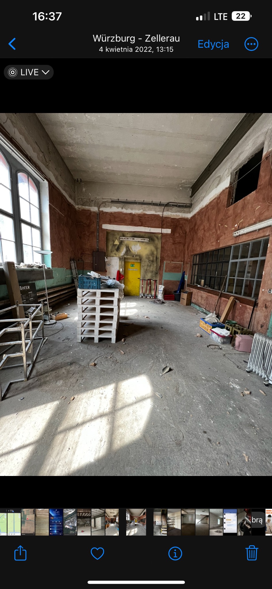 Wnętrze dużego, zaniedbanego pomieszczenia z wysokimi oknami, czerwonymi ścianami z odpadającym tynkiem i żółtymi drzwiami; widoczne palety, skrzynki i resztki materiałów budowlanych.