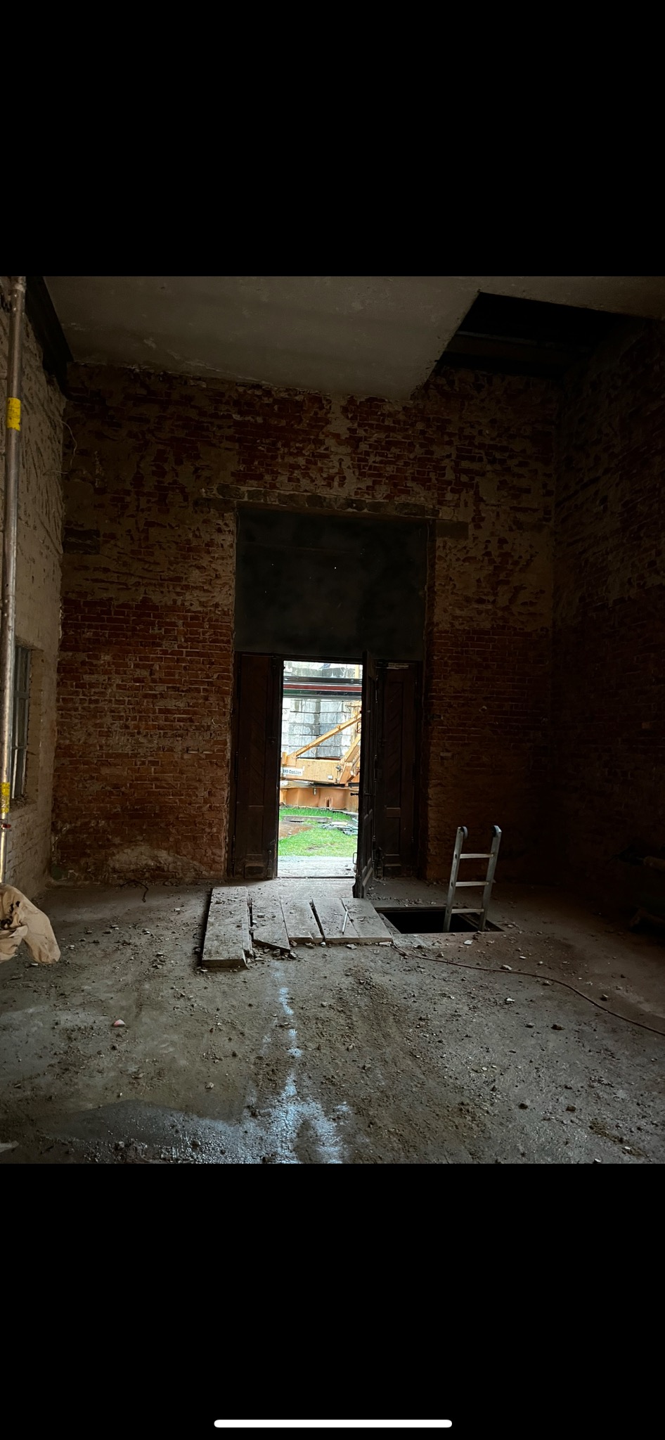 Wnętrze budynku w trakcie rozbiórki, z odsłoniętą cegłą, widocznymi śladami wilgoci na podłodze, otwartymi drzwiami prowadzącymi na zewnątrz i drabinką stojącą przy otworze w podłodze.