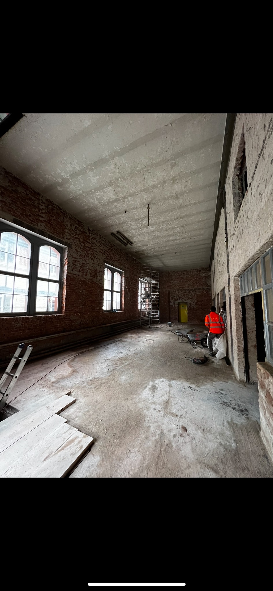 Przestronne wnętrze w trakcie prac demontażowych, widoczne ceglane ściany, surowe wykończenie, drabina oparta o ścianę oraz rusztowanie w głębi pomieszczenia, żółte drzwi w tle.