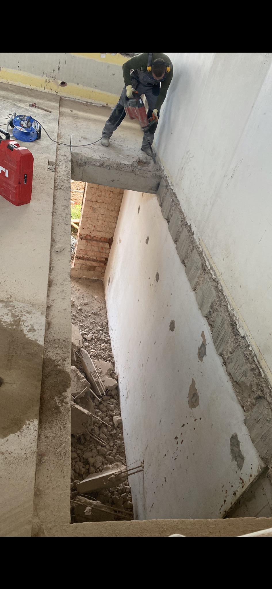 Pracownik w kombinezonie i słuchawkach ochronnych wyburza ścianę za pomocą młota udarowego, widoczne gruz i fragmenty cegieł w otworze.