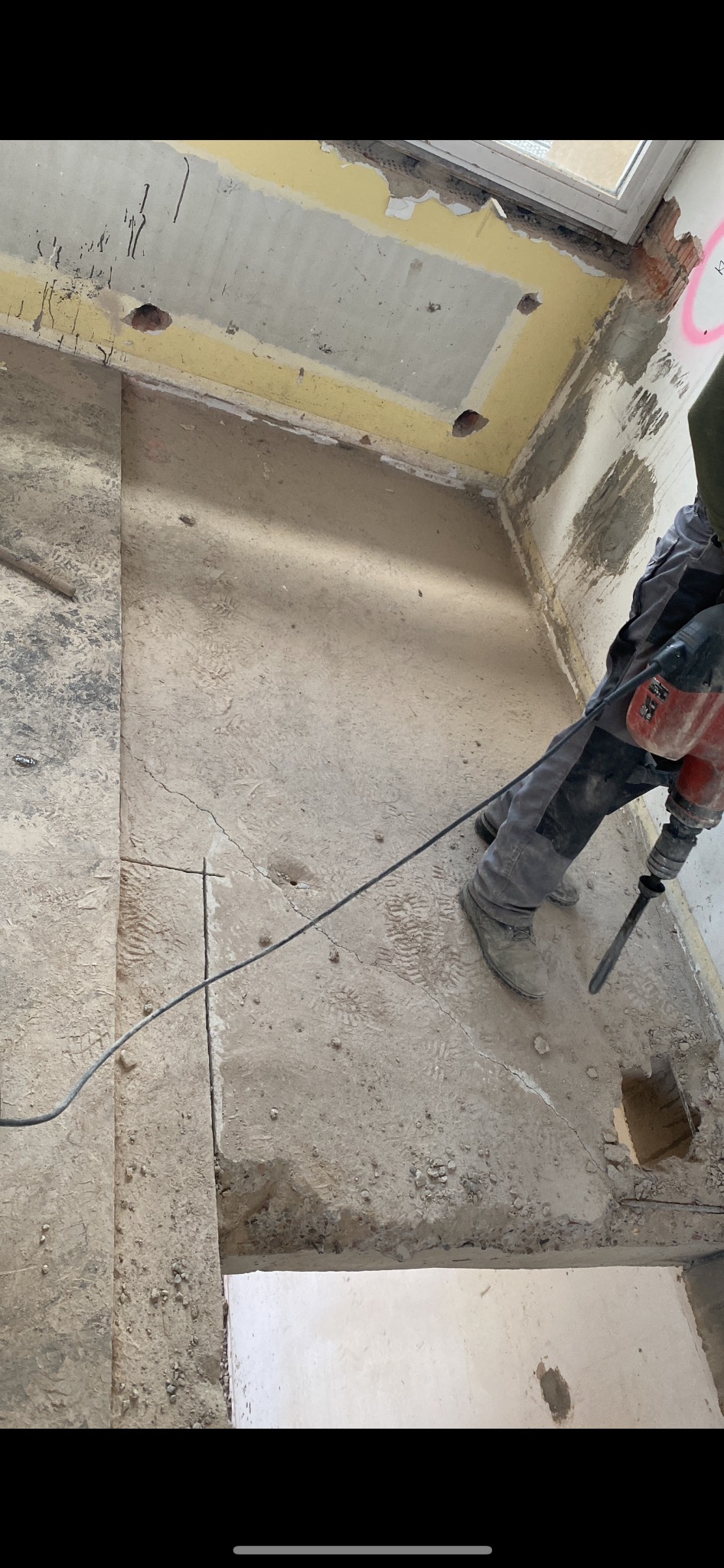 Pracownik w Świdwinie używa młota pneumatycznego do skuwania posadzki w pomieszczeniu przed remontem, widoczne ślady po instalacjach na ścianach i resztki starej farby.