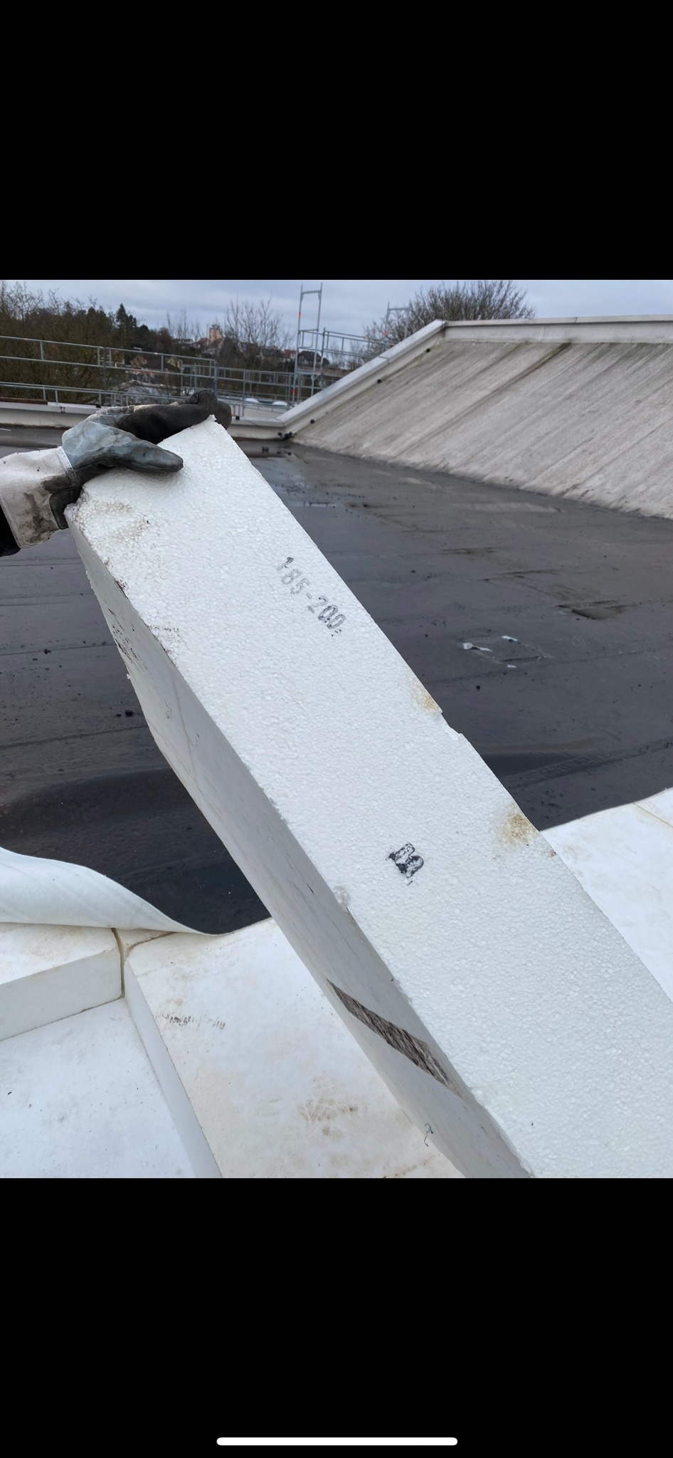 Ocieplenie dachu: pracownik w rękawicy trzyma białą płytę styropianową z nadrukowanym numerem na tle płaskiego, bitumicznego dachu z widocznymi fragmentami ocieplenia i elementami konstrukcyjnymi.