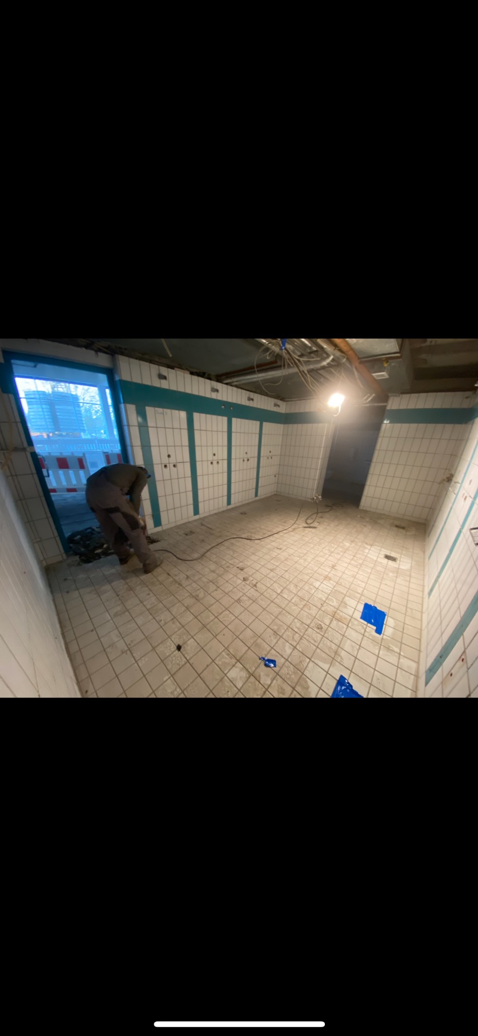 Pracownik w kombinezonie roboczym używa szlifierki do usuwania płytek z podłogi w pomieszczeniu z białymi, kwadratowymi płytkami i zielonymi pasami na ścianach. Widoczne kable i rury pod sufitem.