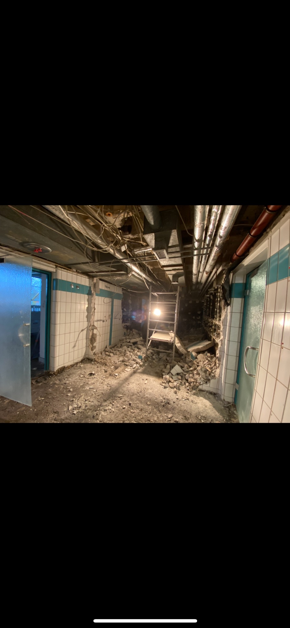 Wnętrze w trakcie wyburzania ściany z białych płytek z turkusowym paskiem, widoczne instalacje pod sufitem i gruz na podłodze, drabina w tle.