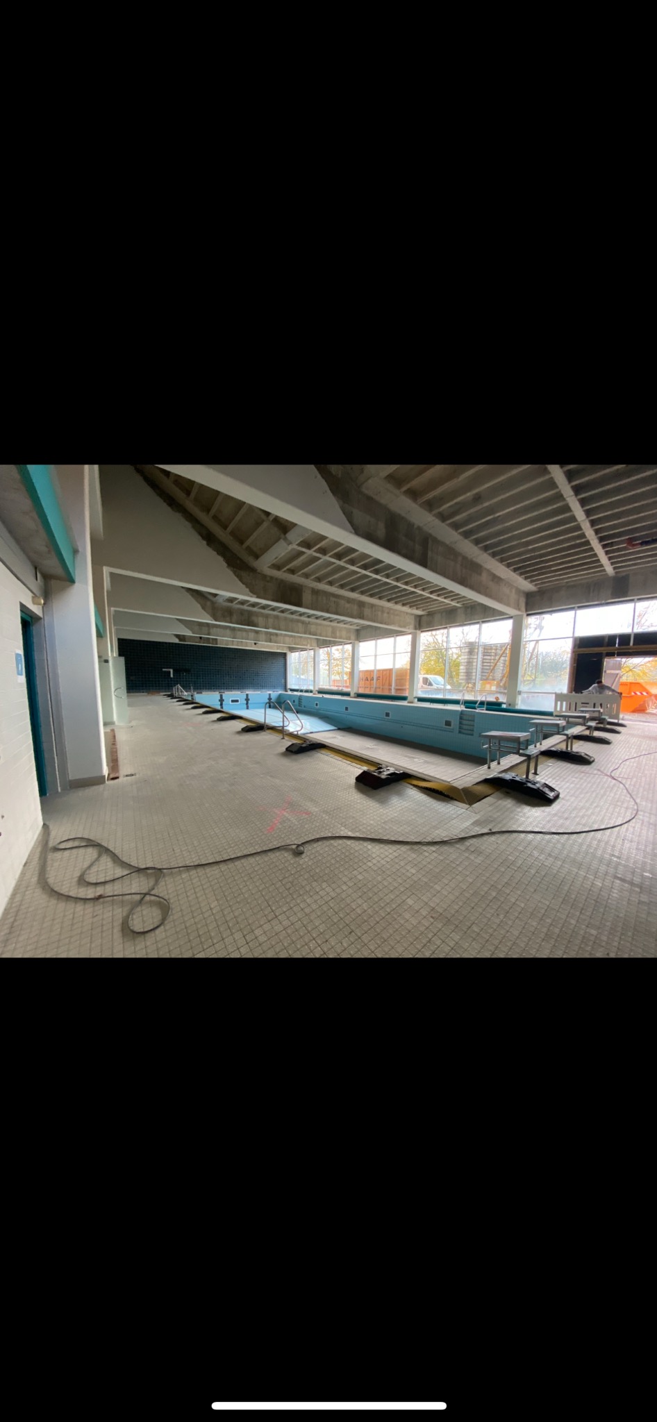 Wnętrze hali basenowej w trakcie remontu: widoczne niebieskie kafelki basenu, rusztowania, kable na podłodze i surowy strop.