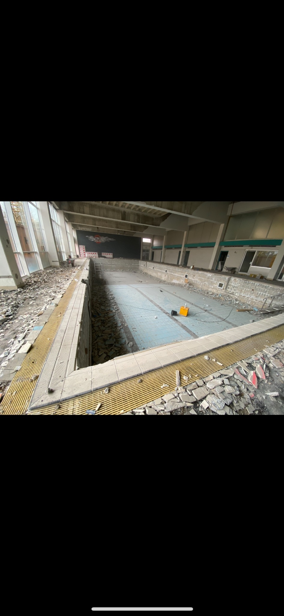 Wnętrze zdemontowanego basenu z częściowo usuniętymi płytkami, widoczne gruz i resztki materiałów budowlanych, żółty reflektor na dnie niecki.