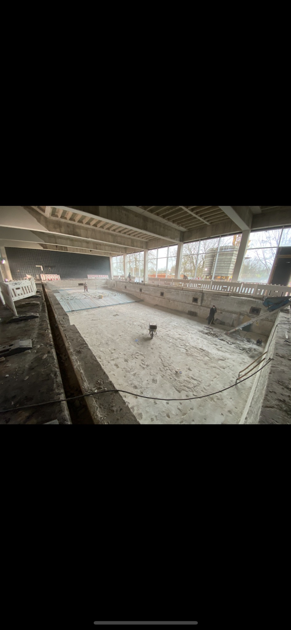 Wnętrze opuszczonego basenu w trakcie rozbiórki, widoczne gruz, narzędzia i pracownicy w oddali.