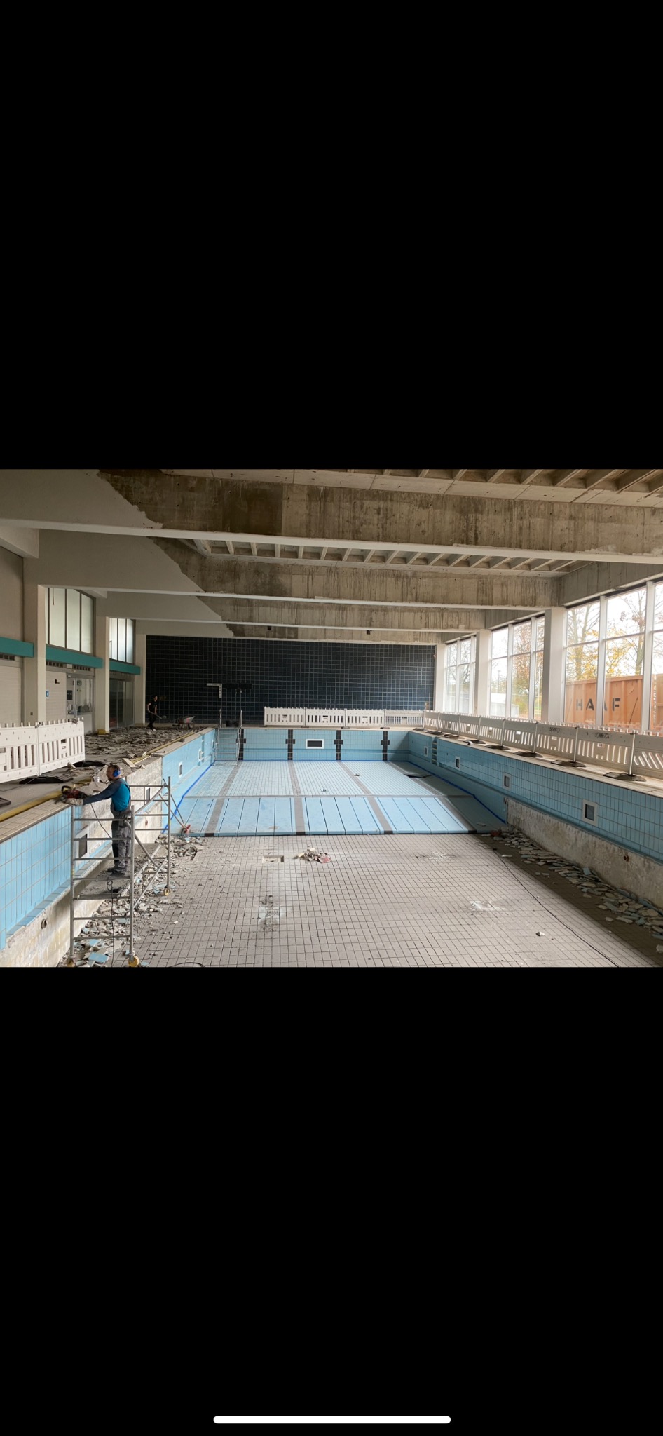 Pracownik w niebieskiej koszuli na rusztowaniu, usuwa płytki ze ściany nieczynnego basenu, widoczne gruz i resztki płytek na podłodze.