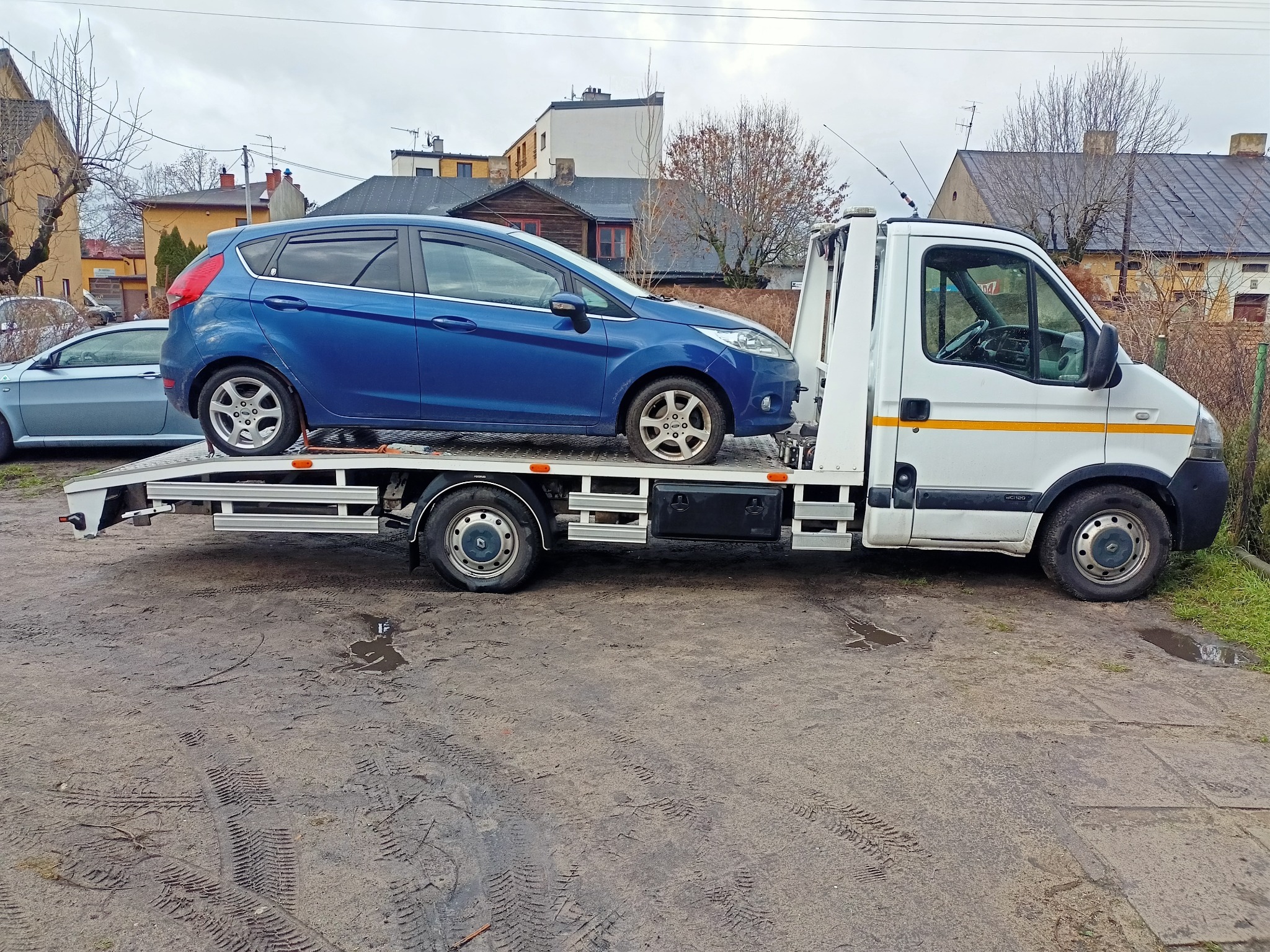Niebieski samochód osobowy załadowany na białą platformę pojazdu transportowego z podnośnikiem, widok z boku na tle zabudowań miejskich i pochmurnego nieba.