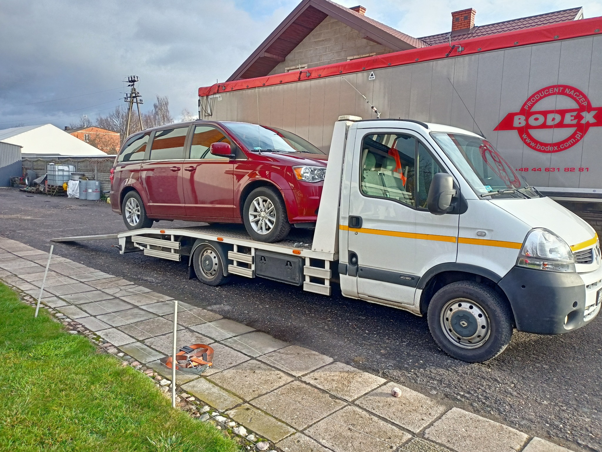 Czerwony minivan na lawecie, biała laweta z żółtymi pasami na tle budynku i naczepy z logo Bodex.
