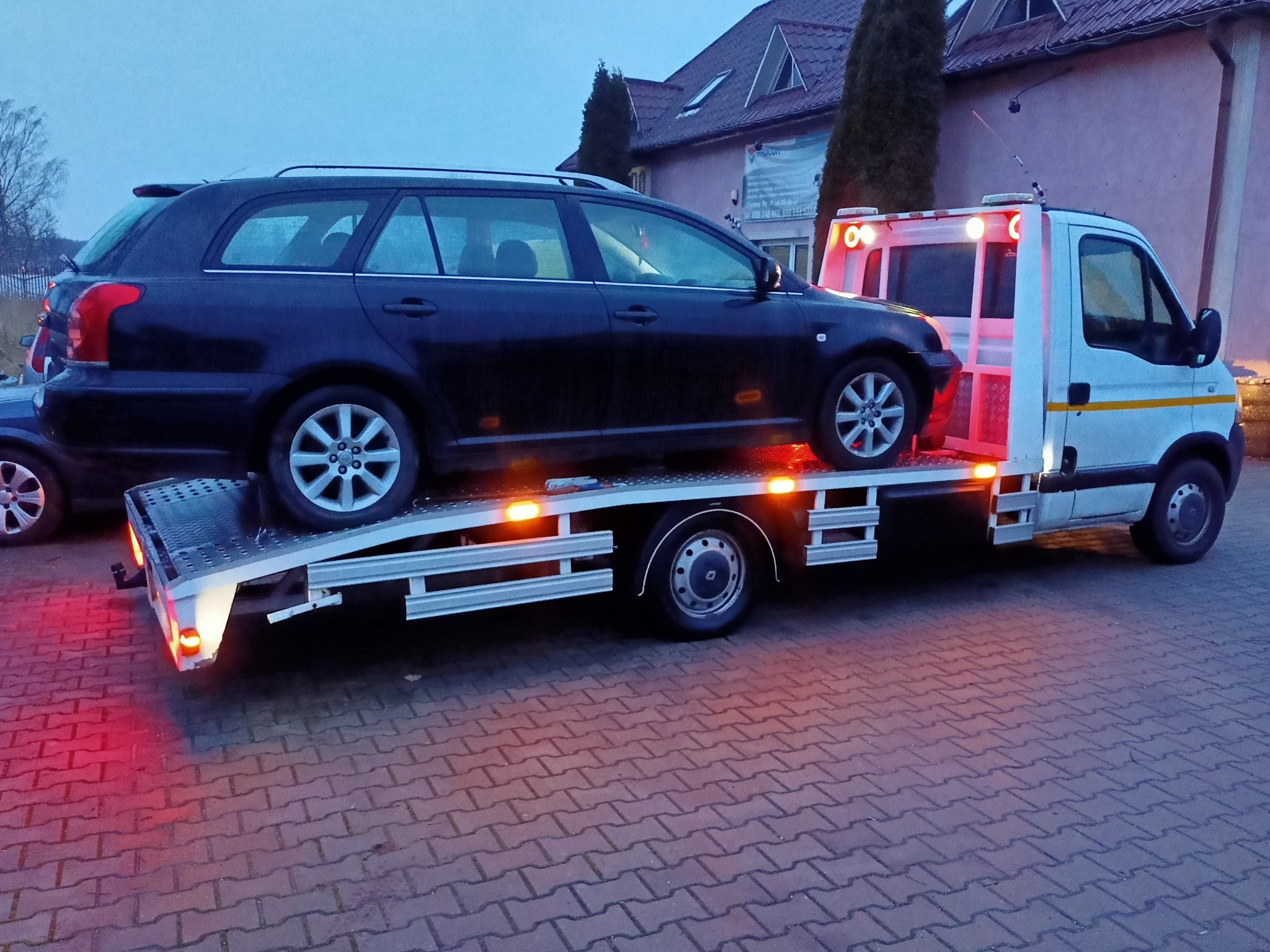 Czarny samochód typu kombi przewożony na platformie transportowej z włączonymi światłami ostrzegawczymi na tle budynku mieszkalnego.