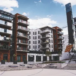 Nowoczesne budynki mieszkalne z cegły i szkła, widoczne balkony z szklanymi balustradami, plac z fontanną i schodami, częściowo zasłonięte przez konstrukcję reklamową.