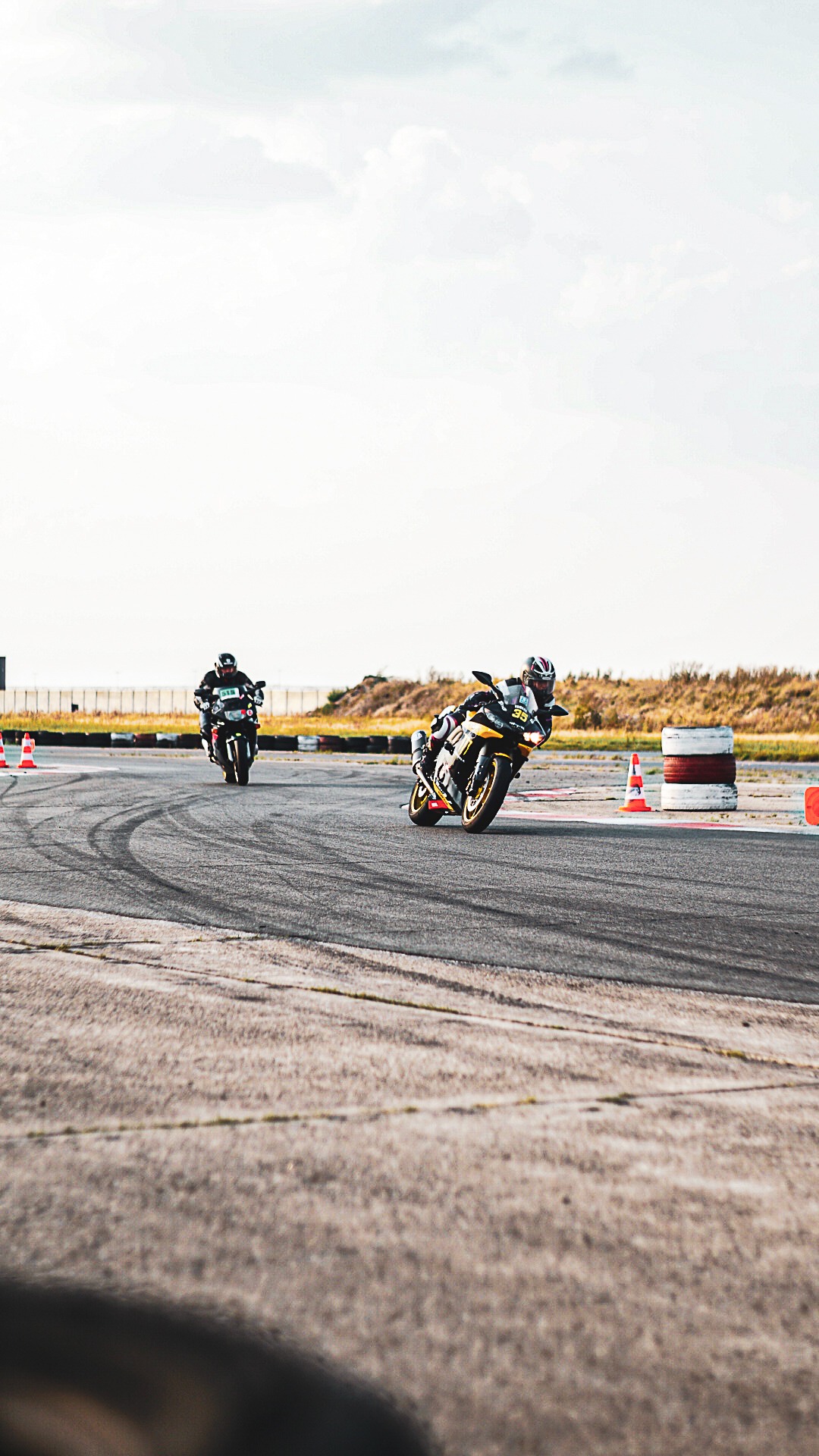 Dwaj motocykliści na torze wyścigowym, jeden w czarno-żółtym stroju pochyla się w zakręcie, drugi w czarnym stroju jedzie prosto w tle, widoczne opony i pomarańczowe pachołki.