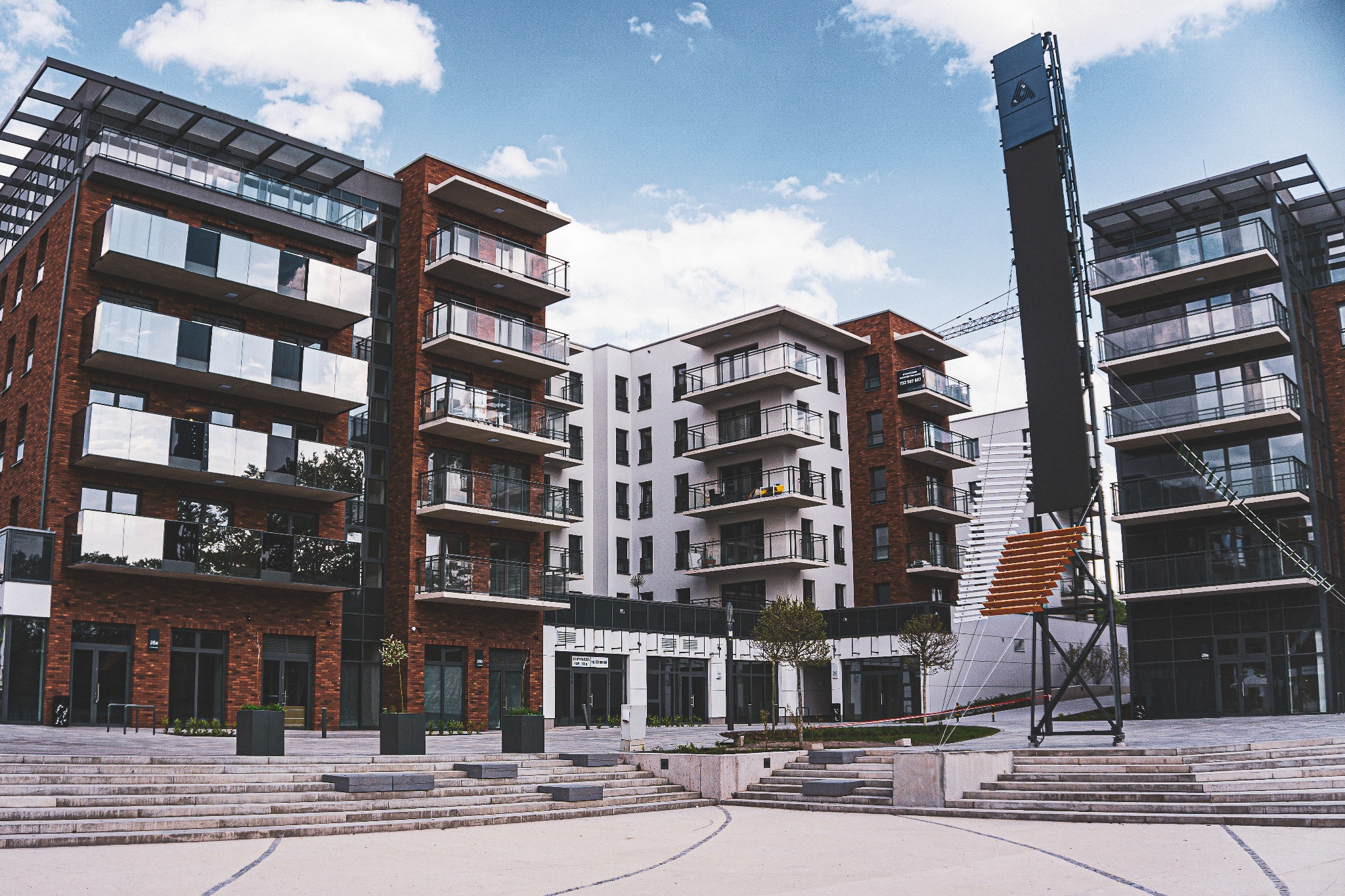 Nowoczesne budynki mieszkalne z cegły i szkła, widoczne balkony z szklanymi balustradami, plac z fontanną i schodami, częściowo zasłonięte przez konstrukcję reklamową.