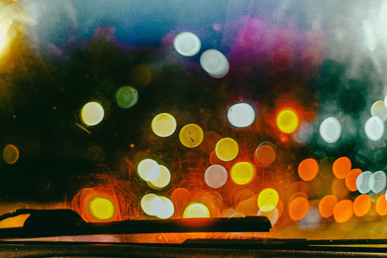 Rozmyte światła miasta widziane przez przednią szybę samochodu, z widocznym wycieraczką w dolnej części kadru.