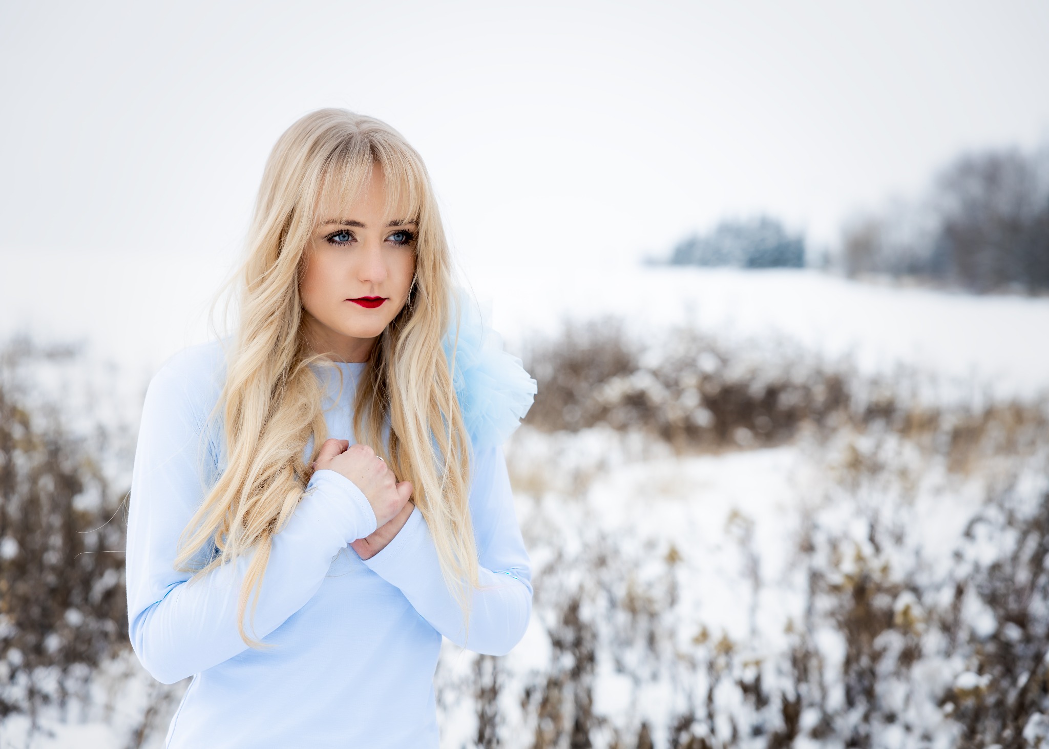 Portret kobiety z blond włosami i czerwonymi ustami w jasnoniebieskiej sukience na tle zimowego krajobrazu z krzakami i drzewami pokrytymi śniegiem.