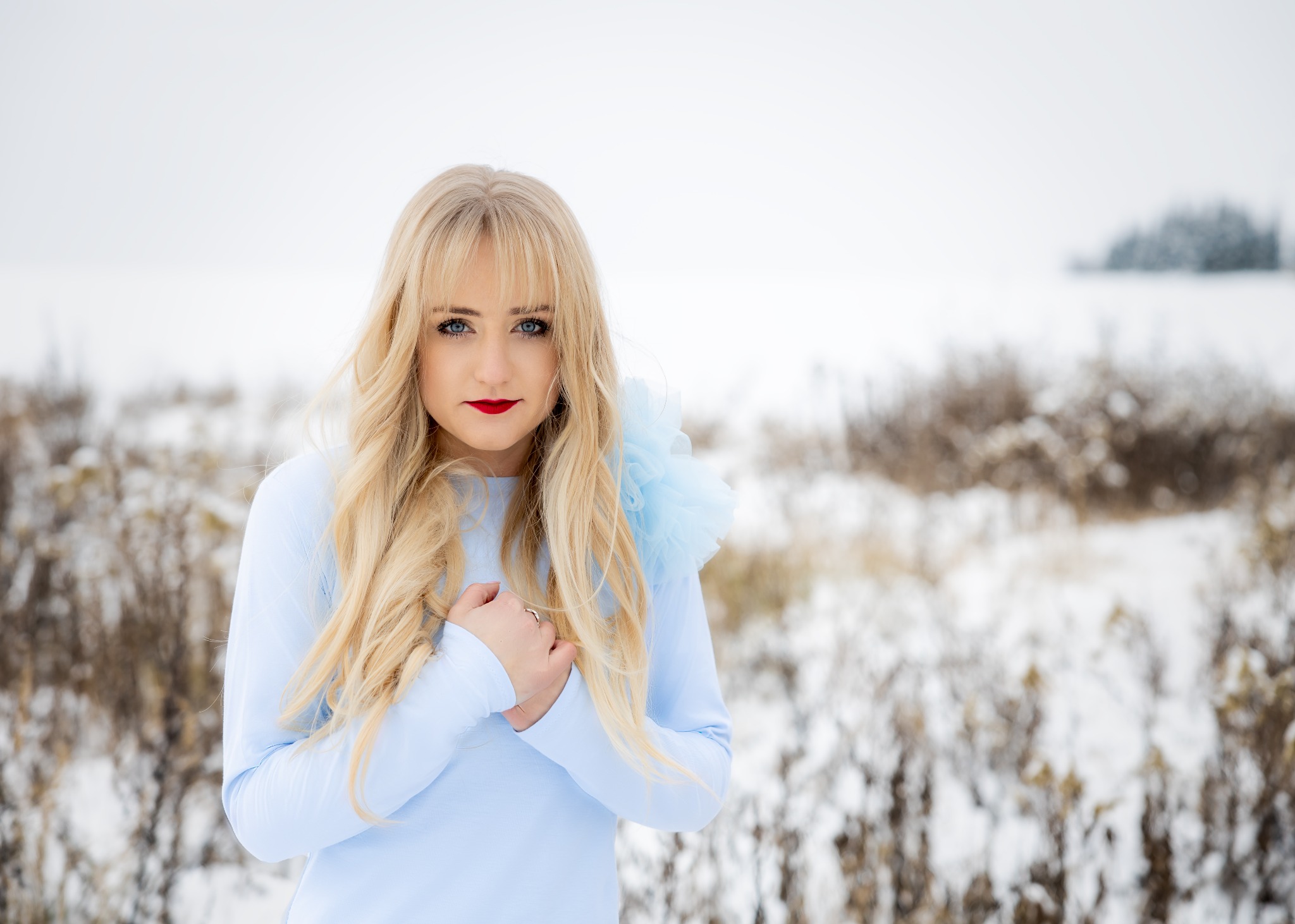 Portret kobiety w błękitnej sukience z tiulowym kołnierzem na tle zimowego krajobrazu z zaspami śniegu i brązowymi trawami.