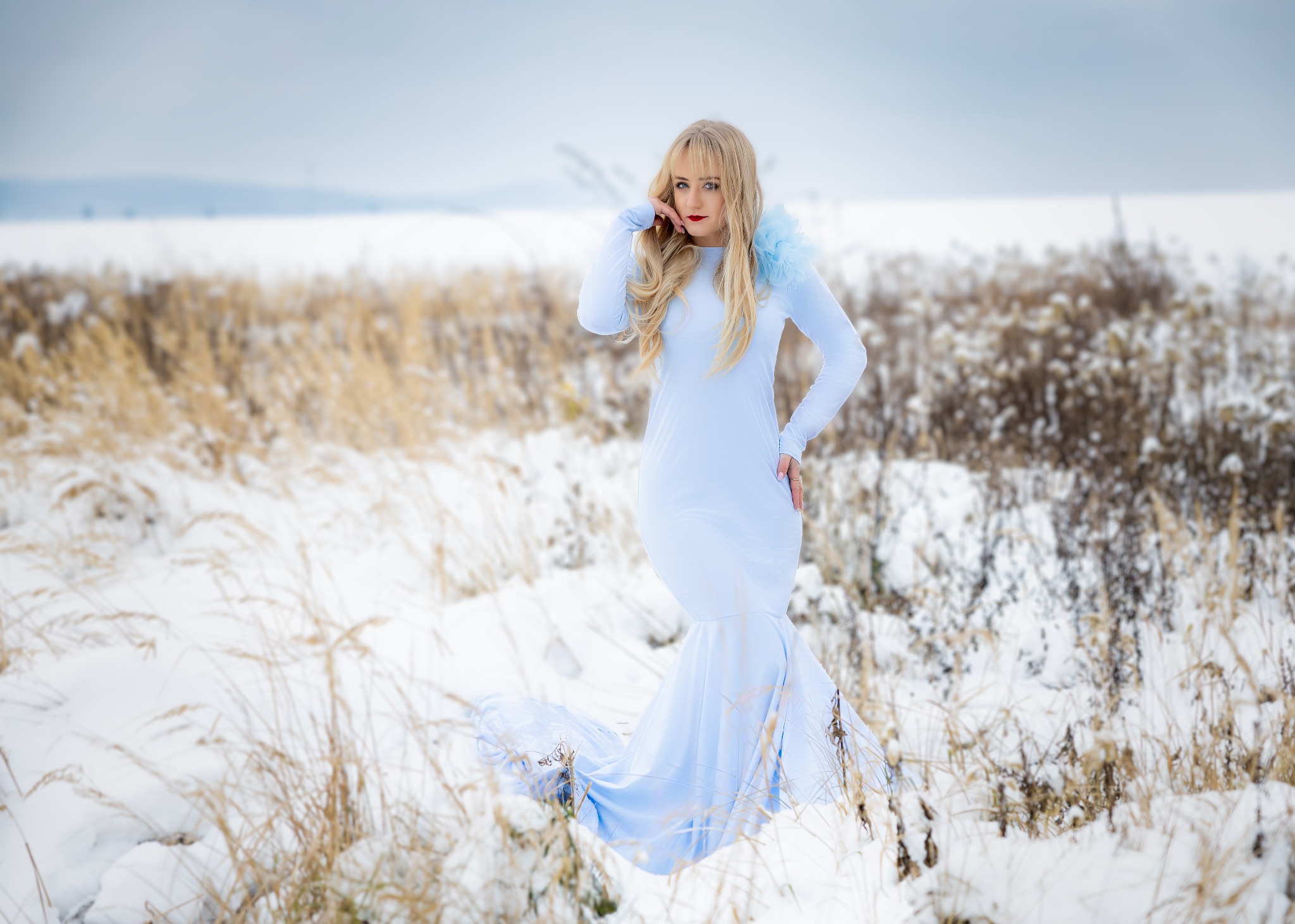 Kobieta w błękitnej sukni pozuje na tle zimowego krajobrazu w Świdnicy, dłoń przy twarzy, delikatny makijaż, długie blond włosy.