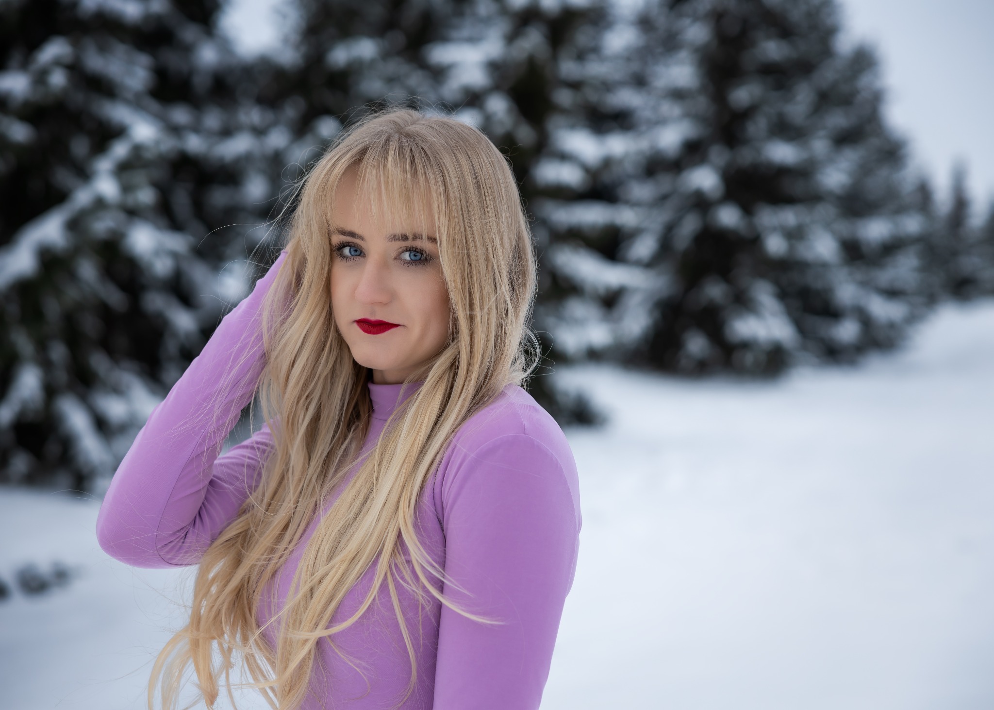 Portret kobiety z blond włosami i czerwonymi ustami w fioletowej bluzce na tle ośnieżonych drzew