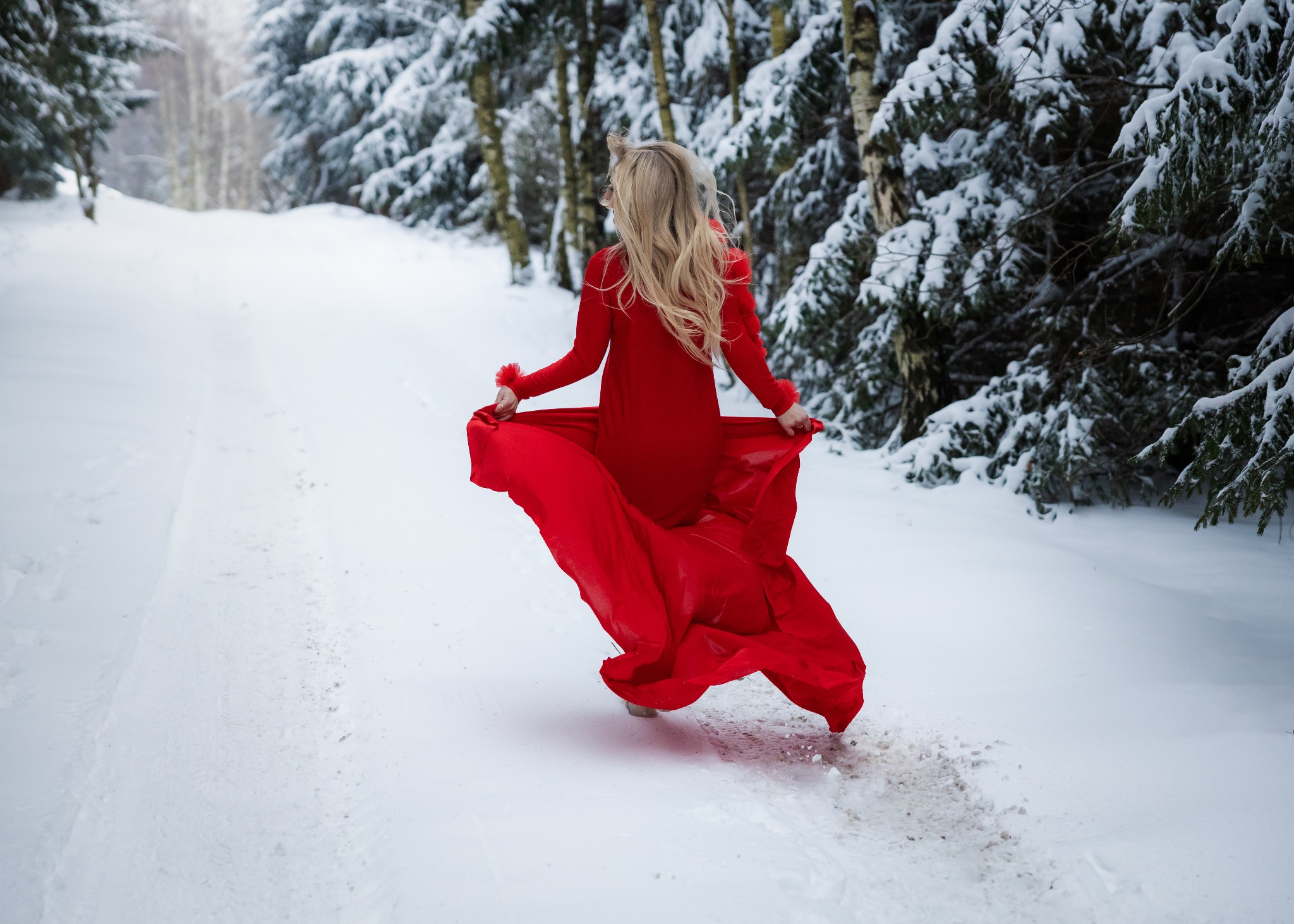 Kobieta w czerwonej sukni biegnie przez ośnieżoną ścieżkę w zimowym lesie, trzymając materiał sukni w dłoniach.