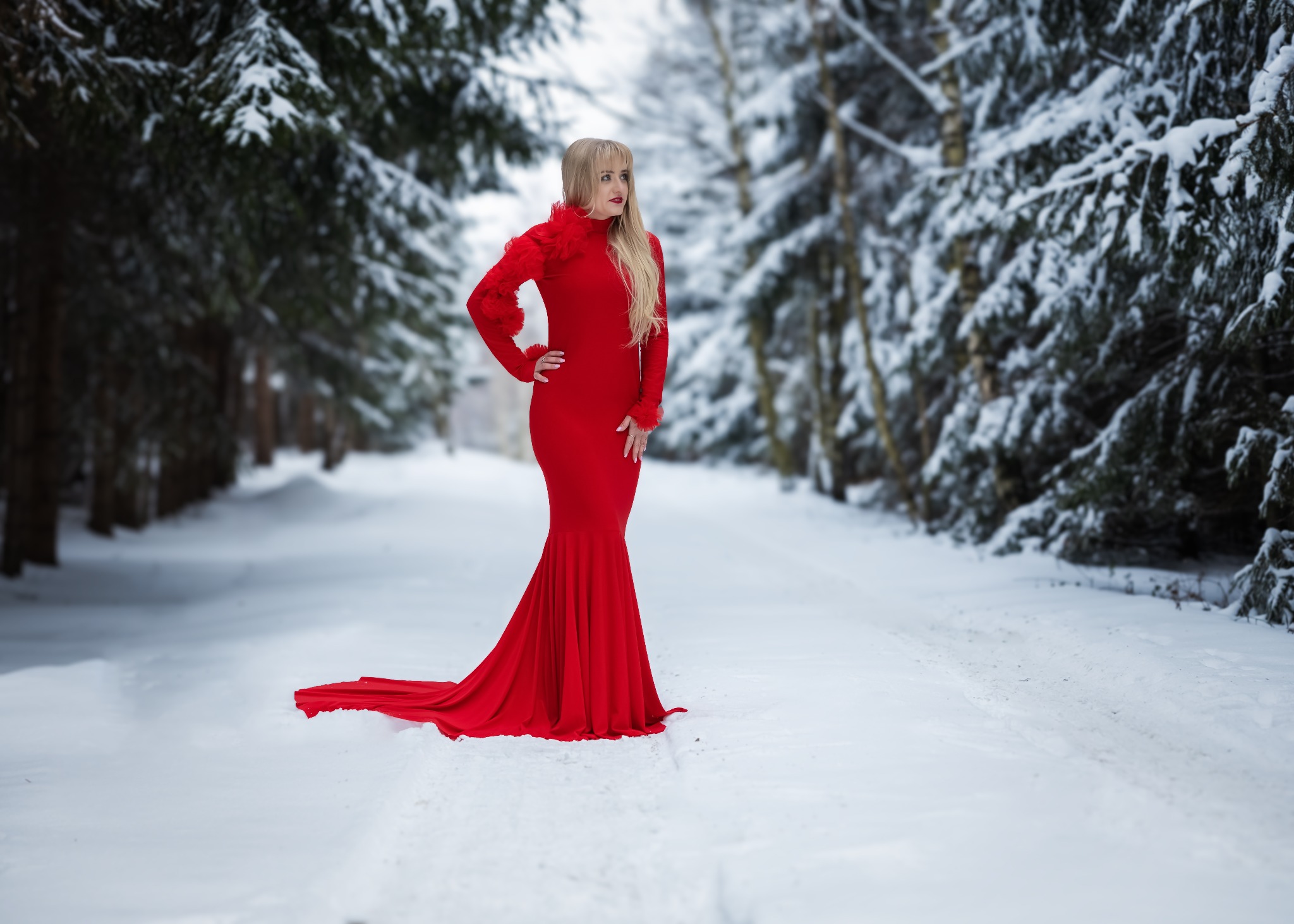 Kobieta w czerwonej, dopasowanej sukni z ozdobnymi elementami pozuje w zimowym lesie, odwrócona w bok, z ręką na biodrze i długimi blond włosami.
