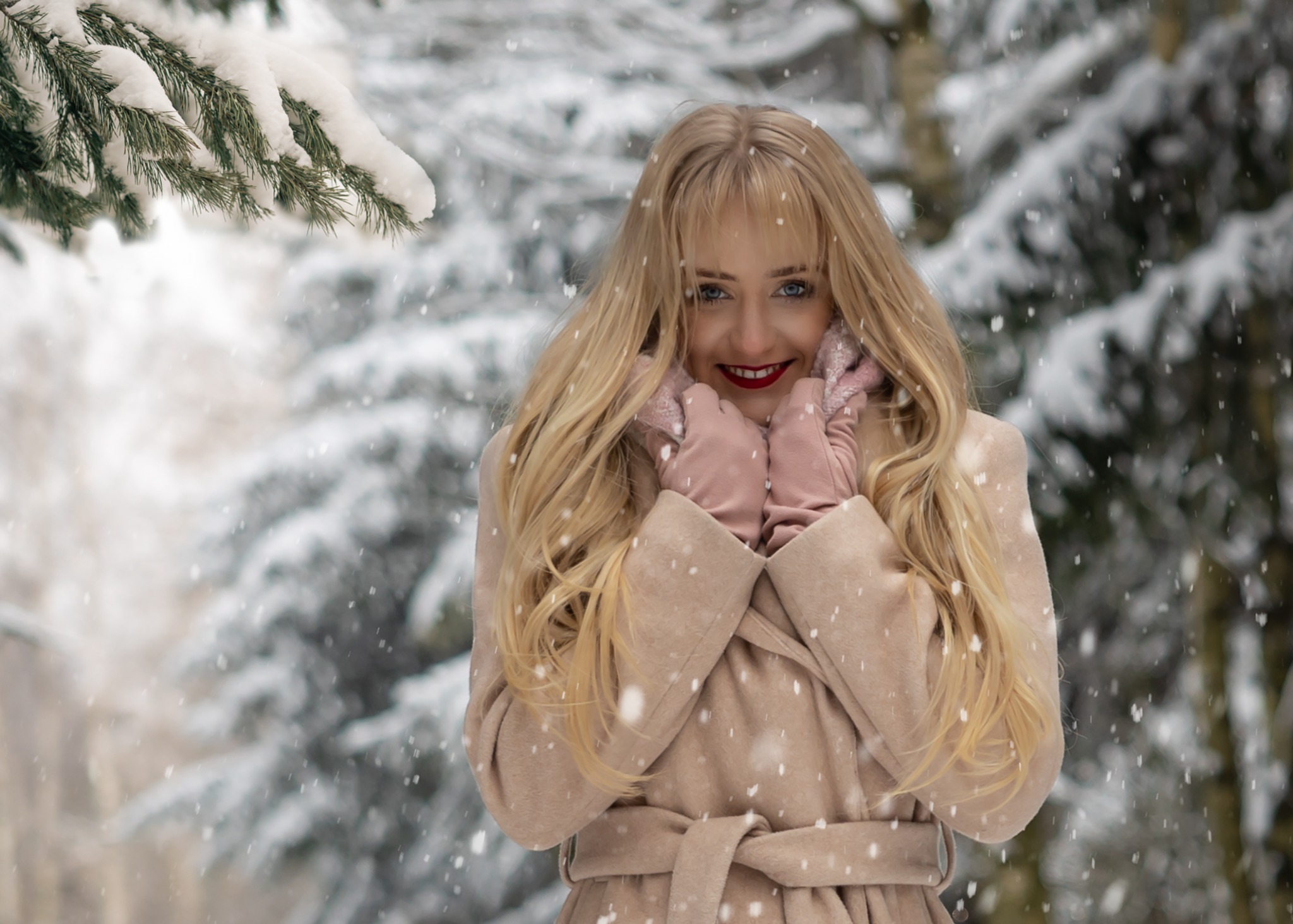 Uśmiechnięta blondynka w beżowym płaszczu i różowych rękawiczkach na tle ośnieżonego lasu podczas opadów śniegu.