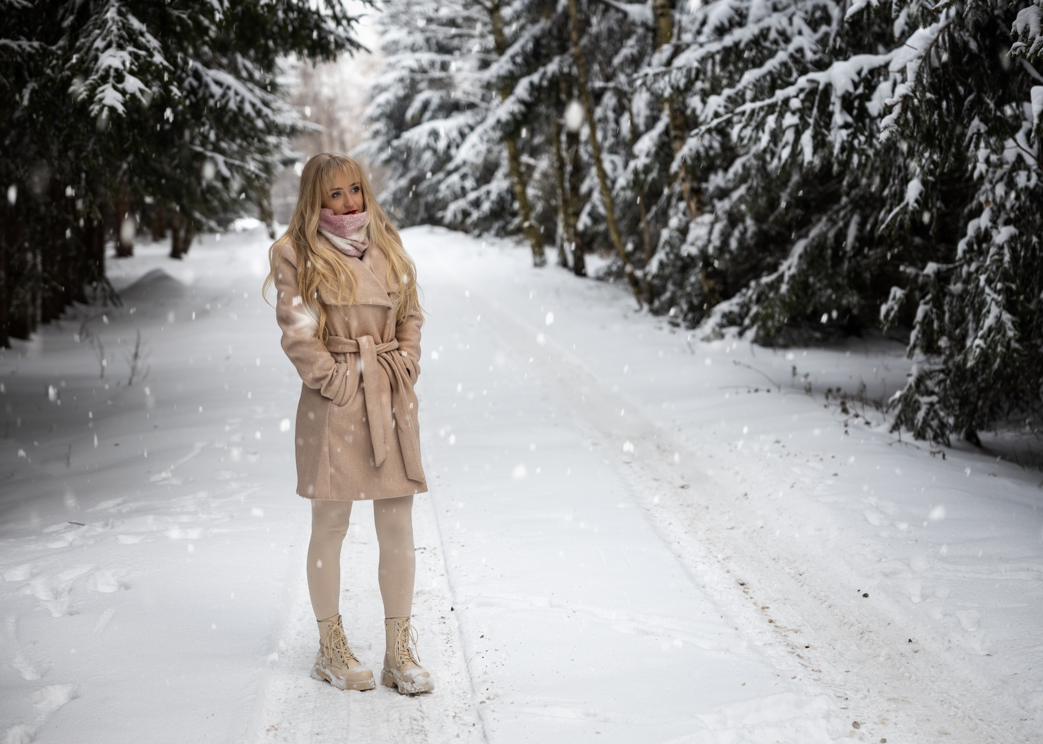 Kobieta w beżowym płaszczu i botkach na śnieżnej drodze w lesie, delikatny uśmiech, padający śnieg.